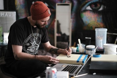 A young man wearing a beanie sitting at his desk with a laptop and drawing on a small surface. Opportunities for creatives.