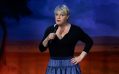 British comedian Eddie Izzard brings 'Eddie Izzard – The Remix Tour Live' to Australia later this year. The photo shows Izzard on stage, posing with a microphone held close to her red-painted lips in her right hand, and her left hand on her hip. She has short blonde hair and wears a long-sleeved black shirt and a ruffled blue skirt.