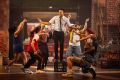A man is standing on a milk crate. There are people dancing around him in a production of In The Heights.