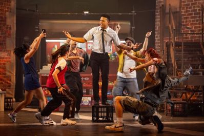A man is standing on a milk crate. There are people dancing around him in a production of In The Heights.