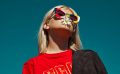 A young blonde woman wearing coloured sunglasses with a diary sticking out of her mouth against a blue sky-like backdrop. Opportunities.