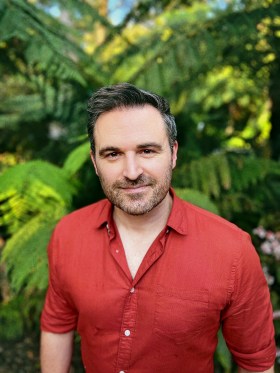 A man in a red shirt standing against foliage. Mark Mupotsa-Russell, author of The Wolf Who Cried Boy.