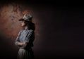 Nina Korbe as Molly Johnson in a promotional image for QPAC and Opera Australia's 2026 co-production, 'The Drover's Wife – The Opera'. The photo shows a First Nations woman standing in profile, her arms folded and her face shadowed beneath a battered Akubra hat. The background is shadowy, highlighting the isolated female figure.