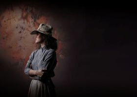 Nina Korbe as Molly Johnson in a promotional image for QPAC and Opera Australia's 2026 co-production, 'The Drover's Wife – The Opera'. The photo shows a First Nations woman standing in profile, her arms folded and her face shadowed beneath a battered Akubra hat. The background is shadowy, highlighting the isolated female figure.