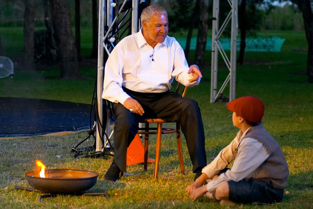 Uncle Noel Tovey in a scene from 2004's 'Skipping on Stars'. The photo shows Uncle Noel, seated on a wooden chair on the grass next to an outdoor stage and speaking sternly to a boy, who wears the clothes of a boy from the turn of the 19th or early 20th centuries.
