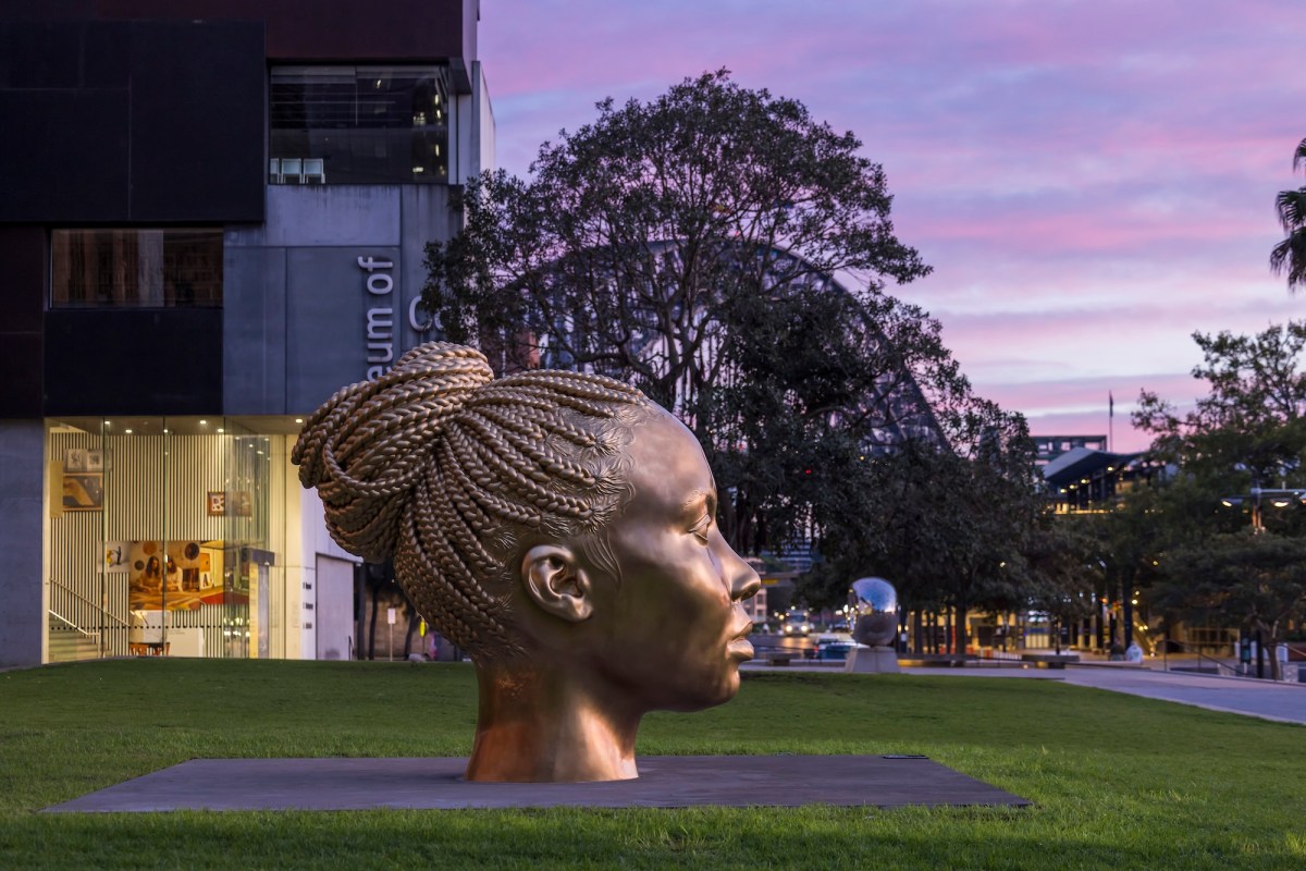 gold sculpture of a black woman's head outside a gallery on lawn at dusk.