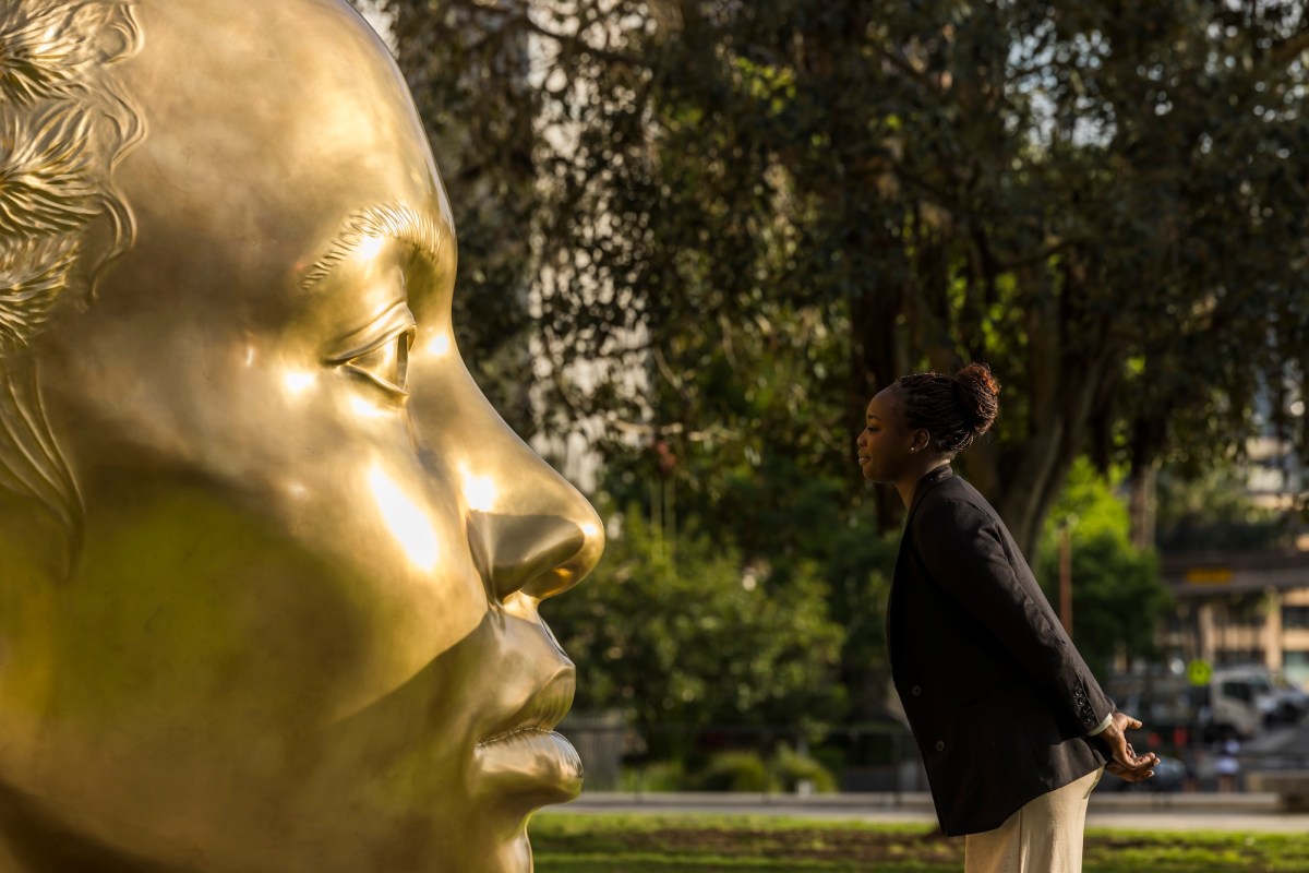 Girl with hair pulled back and dark skin looking at gold sculpture in her likeness.