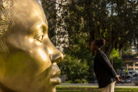 Girl with hair pulled back and dark skin looking at gold sculpture in her likeness.