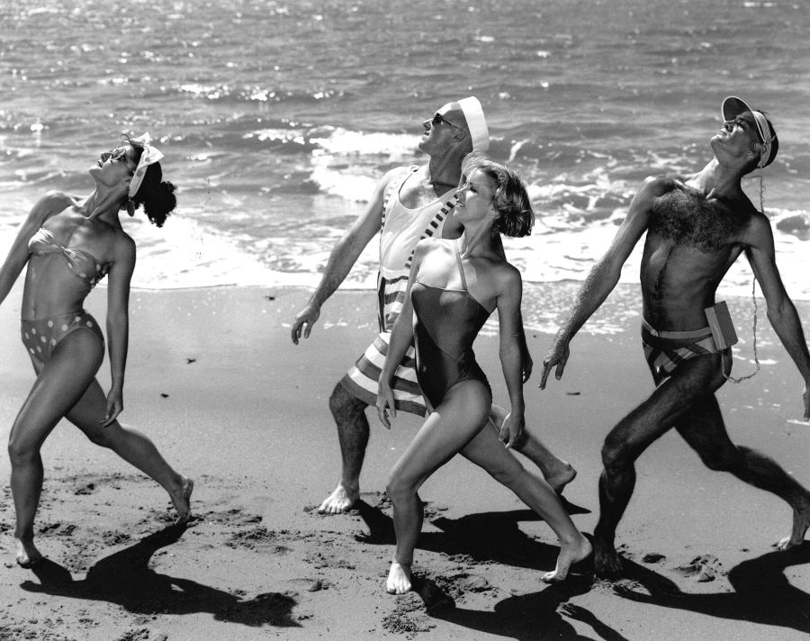 Dancenorth's 1986 work, 'Sun, Sea and Sandwiches'. L-R: Luisa Barisic, Brryn Smith, Suzanne Hurley and William Thomas. A black and white photograph taken on a beach. Four dancers, two women and two men, clad in swimwear, adopt the same pose: arms extended, chest raised, heads leaning back and right leg extended backwards into the sand.