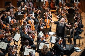 Simone Young conducts 'Mahler Six' with the Sydney Symphony Orchestra in 2018. Photographed from above, Sydney Symphony Orchestra’s Chief Conductor Simone Young - a fair-skinned, blonde haired woman wearing black and standing on a podium with her arms outstretched - conducts the Sydney Symphony. Orchestra members are arrayed around her playing their various instruments.