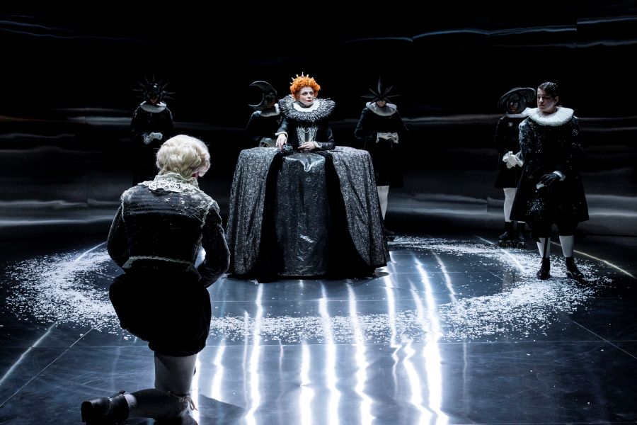 Shannen Alyce Quan as Orlando bows before Amber McMahon's Queen Elizabeth I as Nyx Calder and other cast members look on, in a scene from Belvoir's 2025 production, 'Orlando'. The photo shows Quan, in a blonde wig and Elizbethan garb, kneeling before an imperious queen, who has bright red hair and wears dramatically wide skirts. Other cast members, three in masks and ruffs, one unmasked, look on. A circle of mock-snow is visible on the mirrored stage.