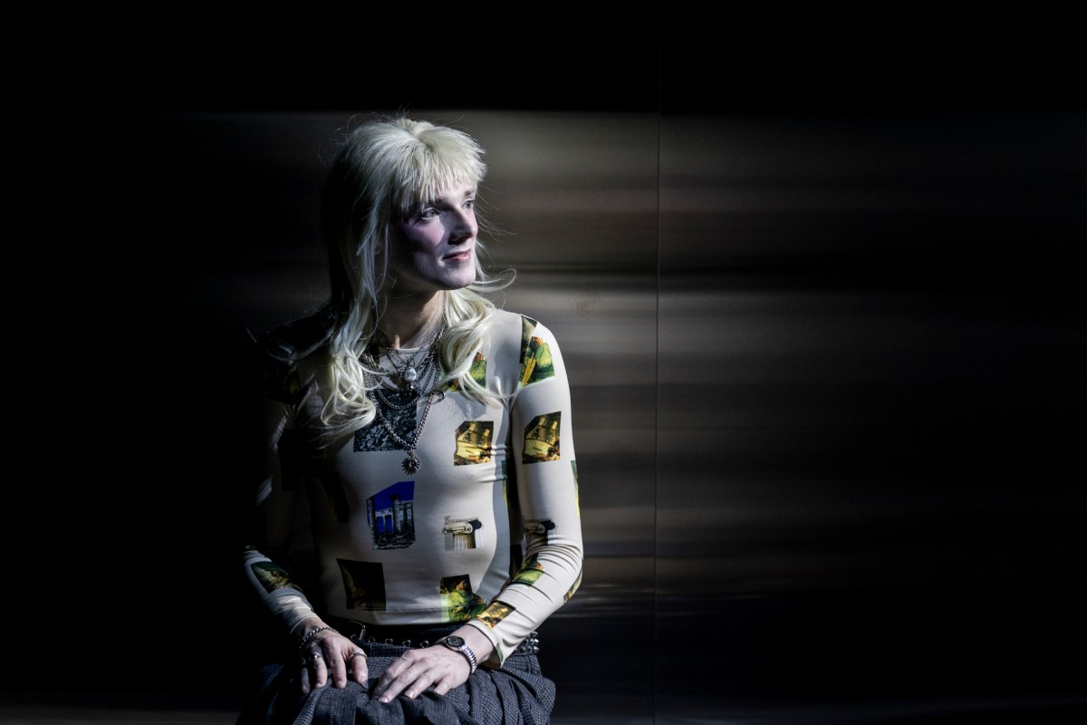 Nic Prior in Belvoir's 2025 production, 'Orlando'. An androgynous figure wearing a punk-inspired blonde wig with a short, spiky fringe and long hair at the back, sits with their hands in their lap against a mirrored background. They are also wearing a pleated grey skirt and skin-hugging white and yellow top.