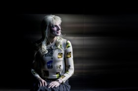 Nic Prior in Belvoir's 2025 production, 'Orlando'. An androgynous figure wearing a punk-inspired blonde wig with a short, spiky fringe and long hair at the back, sits with their hands in their lap against a mirrored background. They are also wearing a pleated grey skirt and skin-hugging white and yellow top.