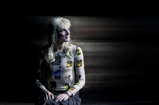Nic Prior in Belvoir's 2025 production, 'Orlando'. An androgynous figure wearing a punk-inspired blonde wig with a short, spiky fringe and long hair at the back, sits with their hands in their lap against a mirrored background. They are also wearing a pleated grey skirt and skin-hugging white and yellow top.