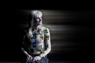 Nic Prior in Belvoir's 2025 production, 'Orlando'. An androgynous figure wearing a punk-inspired blonde wig with a short, spiky fringe and long hair at the back, sits with their hands in their lap against a mirrored background. They are also wearing a pleated grey skirt and skin-hugging white and yellow top.