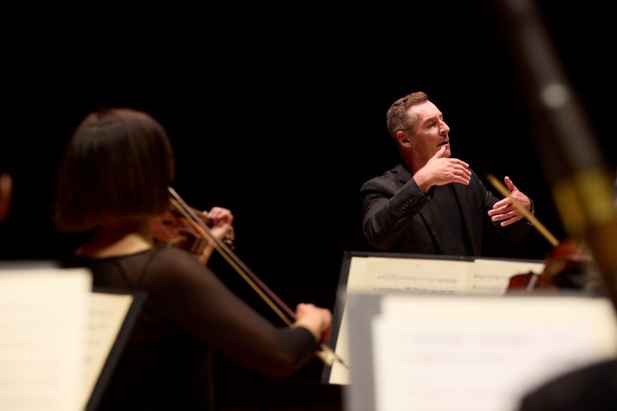 The Australian Brandenburg Orchestra recently launched its 2026 season. The photo depicts the Brandenburg in rehearsal; in the foreground are a slightly blurred series of music stands and scores,; two violinists are visible, and looking towards an older, fair-skinned man who conducts them with his hands, his eyes closed.