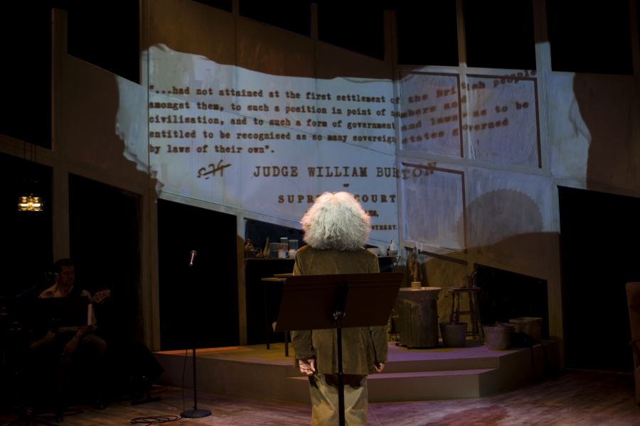 A scene from the 2010 ILBIJERRI and Melbourne Festival co-production,'Jack Charles V The Crown', directed by Rachael Maza. A small Aboriginal man with a great shock of white hair and wearing a brown suit, stands with his back to the camera, looking at a projected document on stage.