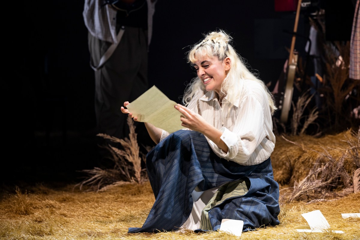 Kala Gare in the MTC's 'My Brilliant Career', 2024. My Brilliant Career the musical is touring in 2026. The photo shows Gare, a fair-skinned woman with blonde hair and weaing a long sleeved white shirt and a dark blue skirt, reading a an unfolded letter and smiling. She is sitting on a pile of straw, denoting the story's rural setting.
