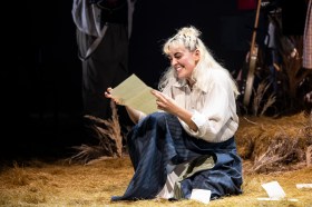 Kala Gare in the MTC's 'My Brilliant Career', 2024. My Brilliant Career the musical is touring in 2026. The photo shows Gare, a fair-skinned woman with blonde hair and weaing a long sleeved white shirt and a dark blue skirt, reading a an unfolded letter and smiling. She is sitting on a pile of straw, denoting the story's rural setting.