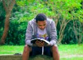 A black man wearing a grey sweater sitting and reading a book in a park. Best new non-fiction.