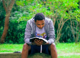 A black man wearing a grey sweater sitting and reading a book in a park. Best new non-fiction.