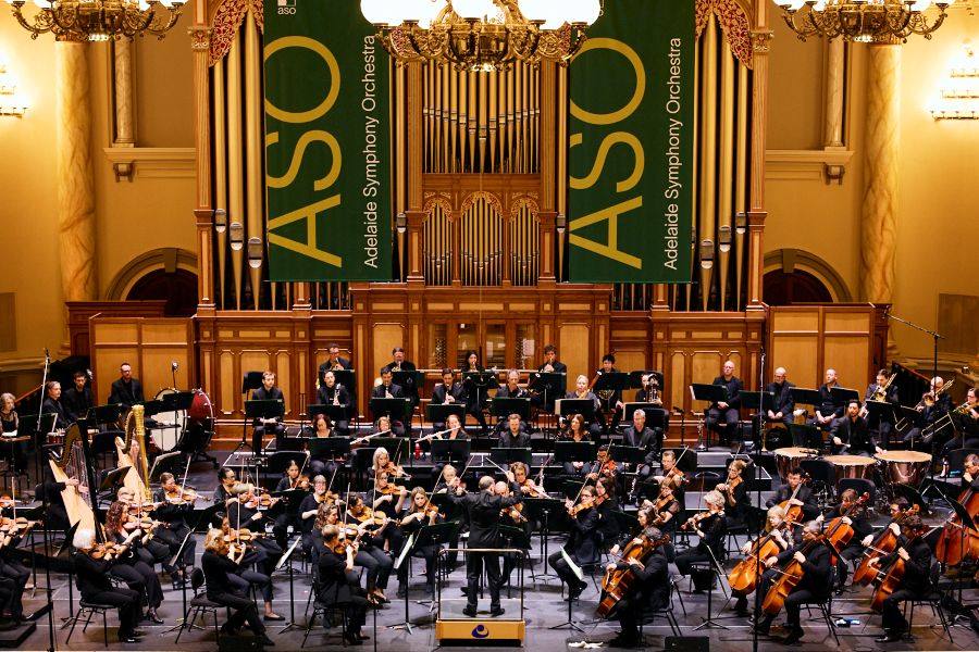 Adelaide Symphony Orchestra performing at Adelaide Town Hall. The photo depicts the many members of the Orchestra clad in formal attire and seated with their instruments on stage. Two banners emblazoned with the letters ASO hang vertically behind them from the Town Hall's pipe organ.