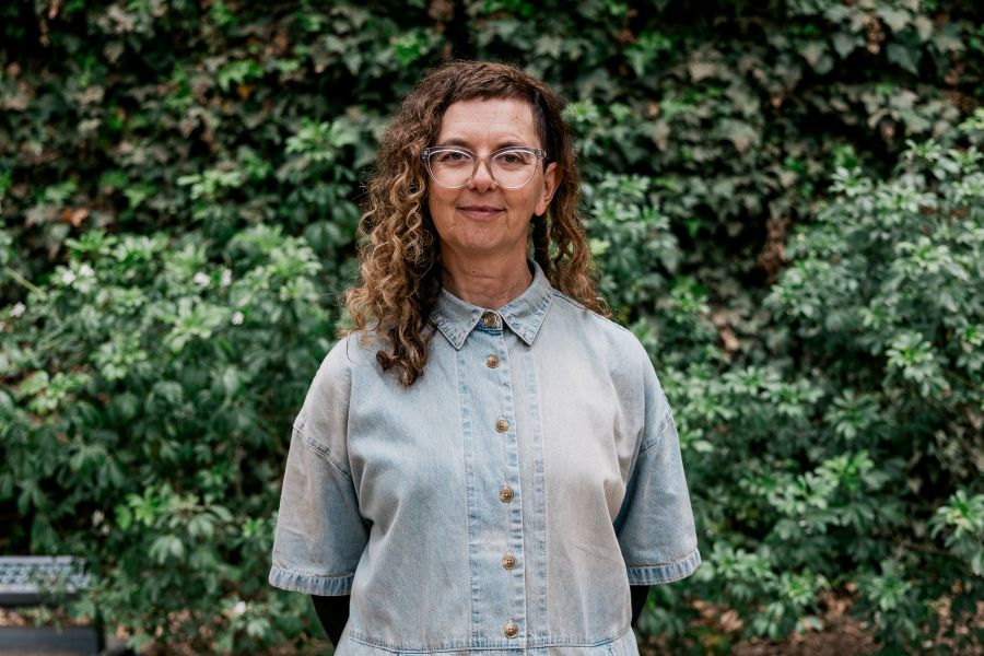 Andrea James is the new Artistic Director and co-CEO of ILBIJERRI Theatre Company. Photo: Jacinta Keefe. James, a Yorta Yorta/Gunaikurnai woman, smiles at the camera with her hands behind her back. She has an ansymmetrical haircut, with long curly brown and blonde hair falling below her right shoulder but cut short on the left, and wears glasses and a pale blue denim shirt buttoned up to her neck.