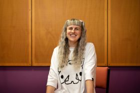 Western Australian composer and producer Elise Reitze-Swensen is a member of the 2025 Minderoo Foundation cohort. The photograph shows Elise Reitze-Swensen, a fair-skinned woman with long blonde hair, smiling at the camera. She wears a long-sleeved white t-shirt decorated with a bold, black-outlined, cartoon-like figure.