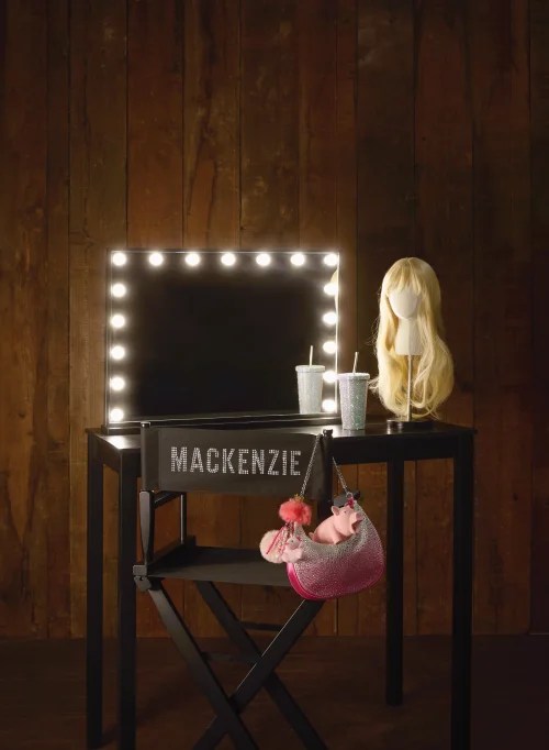 Publicity image for Bell Shakespeare's 2026 season production, 'Mackenzie', a new musical theatre take on 'Macbeth' by Yve Blake. The photo shows a mocked-up dressing room, including a film-set style folding chair with the name 'Mackenzie' written on its back in capital letters, and a make-up mirror ringed by lights. A blonde wig on a wigstand and a disposable cup and straw stand beside the mirror; a garish pink handbag dangles from the chair.