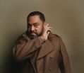 Anthony Taufa, a brown skinned Tongan-Australian actor, poses for the camera with his right hand held up to his left ear, as if trying to hear someone or something - perhaps Banquo's ghost? He wears a heavy brown coat that seems designed to evoke the Scottish cold, and stands against a tan-coloured backdrop.