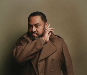 Anthony Taufa, a brown skinned Tongan-Australian actor, poses for the camera with his right hand held up to his left ear, as if trying to hear someone or something - perhaps Banquo's ghost? He wears a heavy brown coat that seems designed to evoke the Scottish cold, and stands against a tan-coloured backdrop.