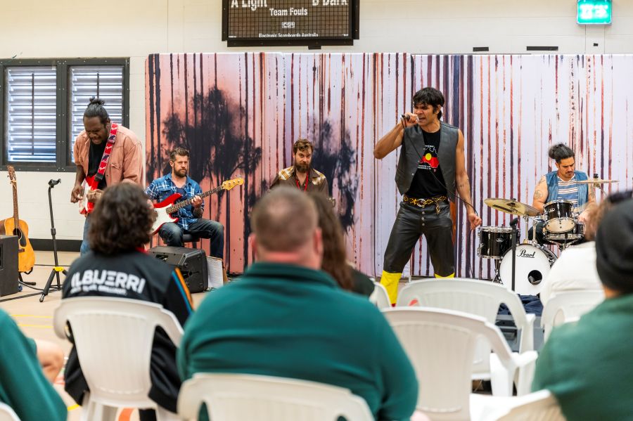 A scene from ILBIJERRI's 'Big Name, No Blankets' Prison Tour; Rachael Maza directed the production. The photograph shows a group of men in green jump suits seated with their backs to the camera, and watching a live band of Aboriginal and non-Aboriginal musicians perform as Warumpi Band.