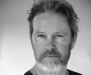 A recent 'Cluedo' publicity photo of actor Craig McLachlan. The black and white photo depicts a middle aged, fair skinned man with a neat grey beard and wearing a black t-shirt.