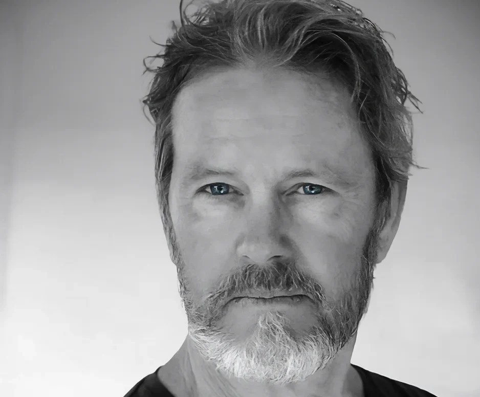 A recent 'Cluedo' publicity photo of actor Craig McLachlan. The black and white photo depicts a middle aged, fair skinned man with a neat grey beard and wearing a black t-shirt.