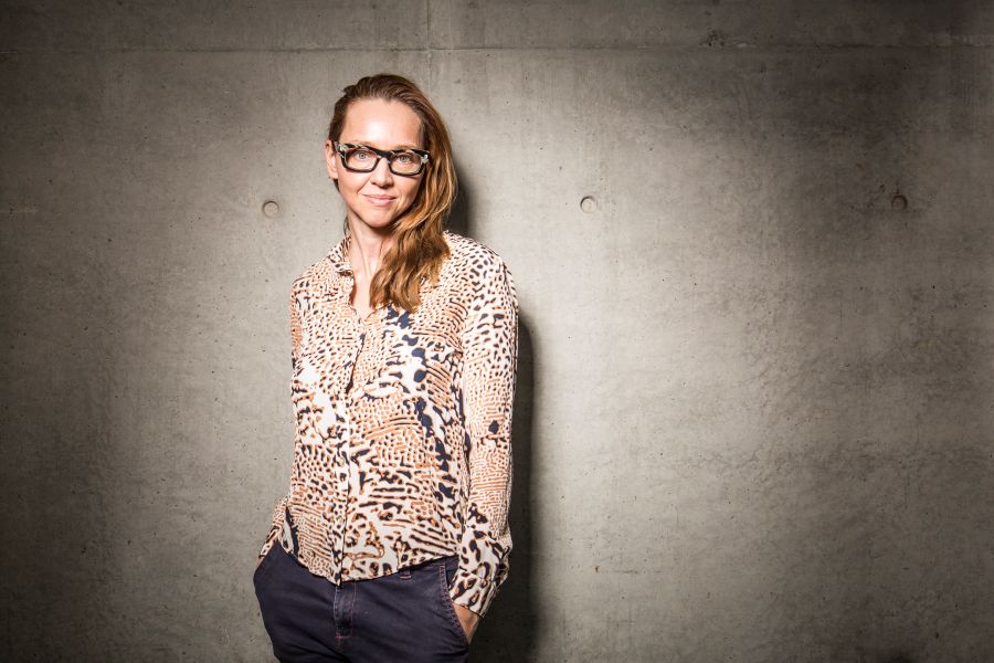 Danielle Micich finishes up at Force Majeure in December 2025. The photo shows Micich, a fair-skinned woman with red hair swept over her left shoulder and wearing glasses and a brown and black spotted top (reminiscent of a leopard or giraffe skin) posing against a grey concrete background. 