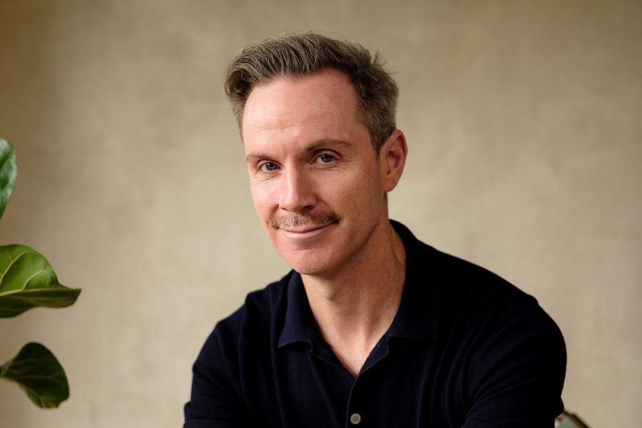 Dean Bryant in the Malthouse Theatre's new Artistic Director. The photograph depicts a fair-skinned man with short brown hair and a neatly trimmed moustache, smiling at the camera. He wears a black collared shirt open at the neck, and sits in front of neutral grey-brown background.