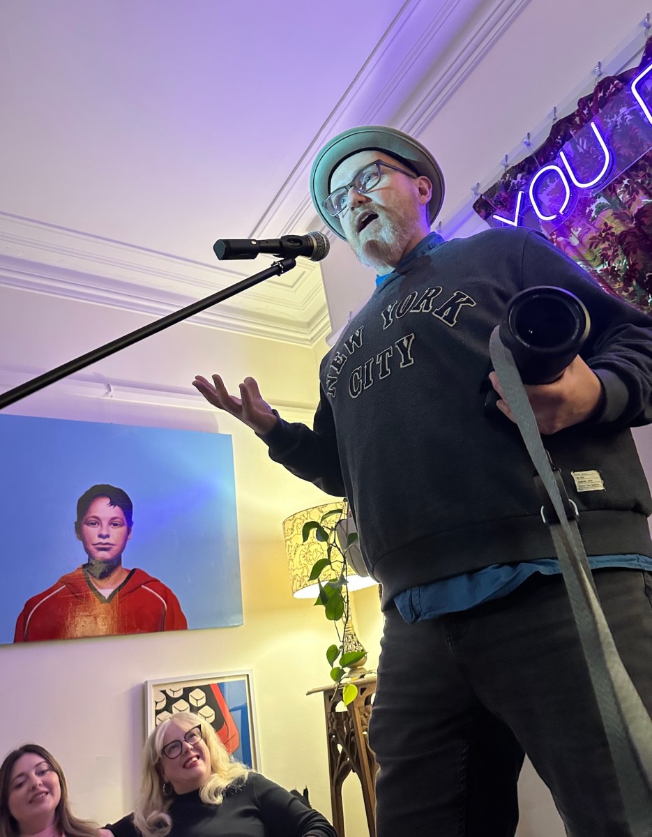 Man with hat on and glasses at microphone in lounge room concert. House concerts