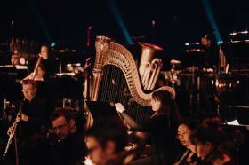 Adelaide Symphony Orchestra performs in the Adelaide Festival Centre's Festival Theatre. An atmospheric photo of Orchestra members on a darkened stage: a dark-haired, fair-skinned woman wearing glasses and playing a large, standing harp is the focal point of the image.