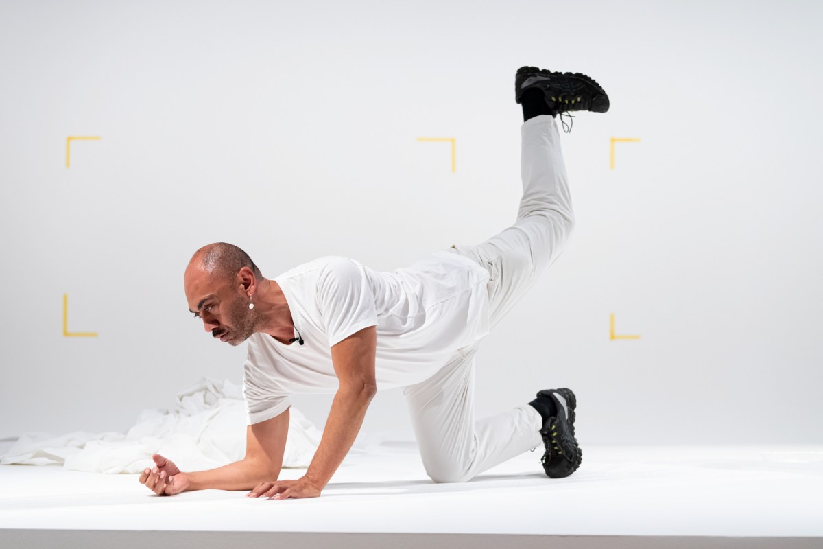 man dressed in white doing balance on hands and knees in art gallery. 
