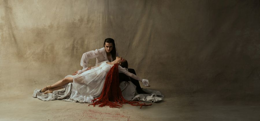 Jurgen Rahimi and Nikki Blain in the West Australian Ballet's 'Dracula', programmed in the company's 2026 season. A pale-skinned man with long black hair and wearing a white shirt, crouches over a supine woman who is also dressed in white. A tangle of red wool is drapped over her left shoulder, representing a gout of blood from her neck.