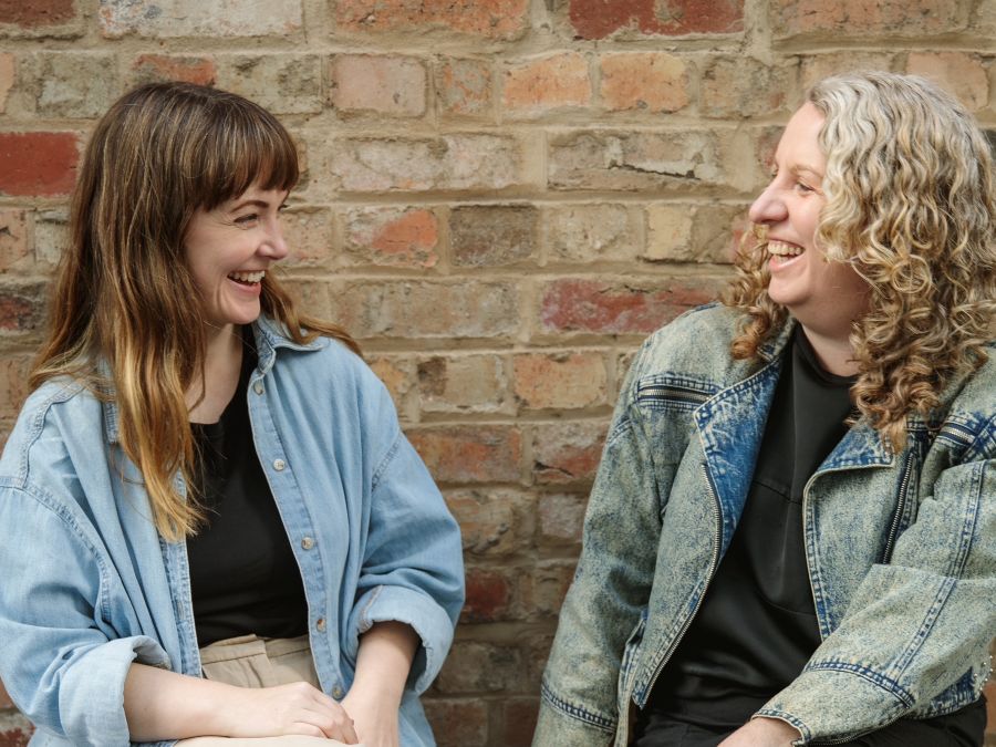 L-R: Keziah Warner and Bron Batten take on salaried roles as Associate Artists at Geelong Arts Centre. The photograph depicts Warner, a fair skinned woman with long brown hair, and Batten, a fair skinned woman with curly blonde hair, sitting opposite one another and laughing.