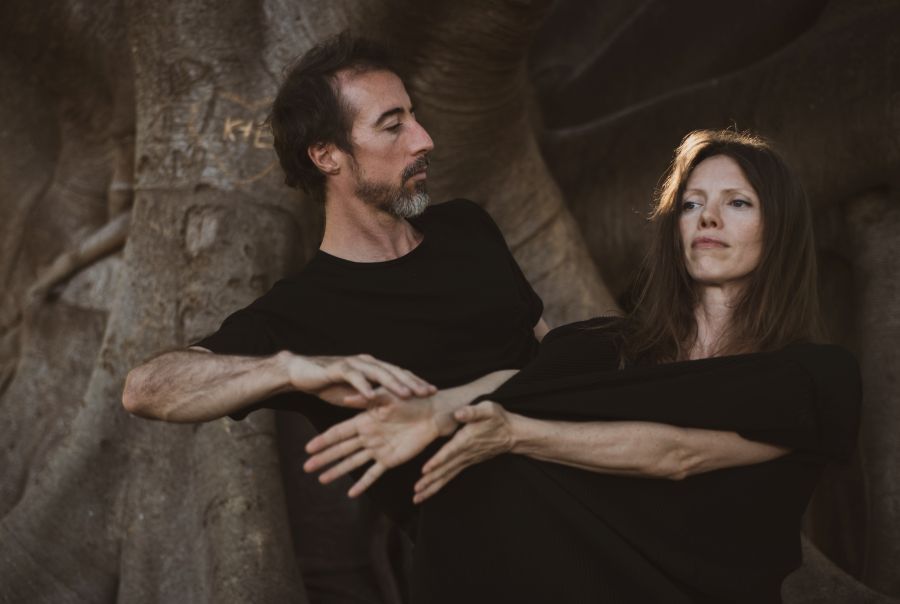 Dancenorth Artistic Director Kyle Page and Associate Artistic Director Amber Haines. The pair, both dressed in black, stand close together in dance poses, arms extended, before the trunk of a large tree.