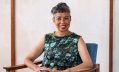 Michelle de Kretsrer's novel 'Theory & Practice' has won the Prime Minister's Literary Award 2025 for Fiction. The photo shows de Kretsrer, an older Asian-Australian woman, sitting in an armchair with her hands in her lap. She is wearing a sleeveless dress depicting a large tree against the blue sky, and has short, greying hair.