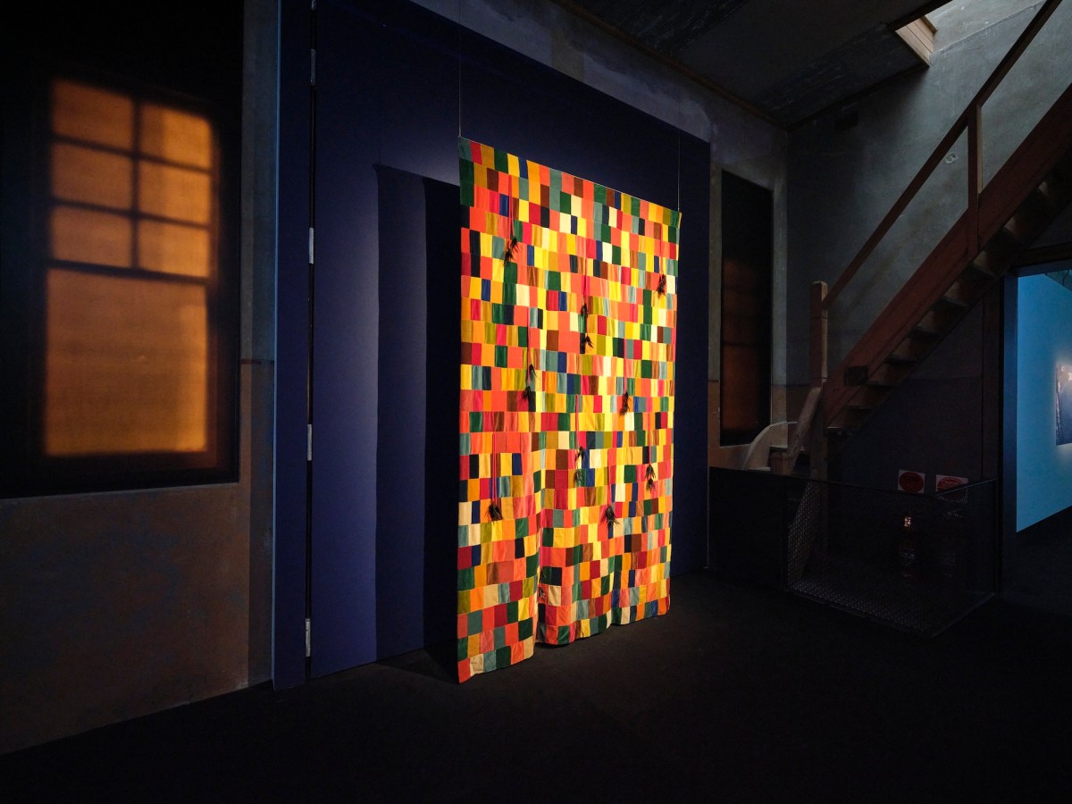 Dimly lit gallery showing a quilt in yellow and red colors. Lisa Reihana
