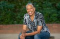 ILBIJERRI's Artistic Director of almost 18 years, Dr Rachael Maza AM. The photo shows Maza, an older Aboriginal and Torres Strait Islander woman with greying hair, sitting in a leafy courtyard. She is smiling, almost laughing, and leans forward with her hands clasped and her elbows on her knees.