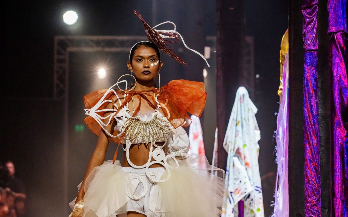 A young First Nations woman wearing an elaborate outfit of earthy tones, featuring bold lines and weaved forms, walking down a runway. Opportunities.