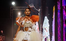 A young First Nations woman wearing an elaborate outfit of earthy tones, featuring bold lines and weaved forms, walking down a runway. Opportunities.