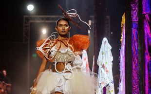 A young First Nations woman wearing an elaborate outfit of earthy tones, featuring bold lines and weaved forms, walking down a runway. Opportunities.