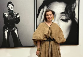 Women with dark hair wearing beige dress standing in front of black and white photographic portrait. Tina Arena