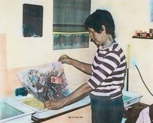 Miriam Stannage: an old looking coloured photograph of a woman at a kitchen holding a printed work on paper she has created.